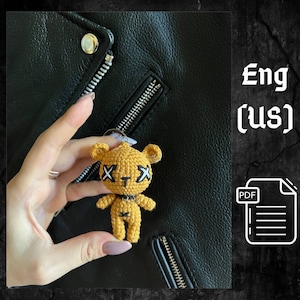May include: A crocheted yellow teddy bear keychain with black X's for eyes and a black choker. The bear is being held by a hand in front of a black leather jacket.