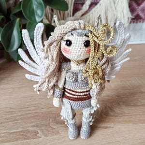 Crocheted valkyrie doll in white wings and armor, standing on wooden table