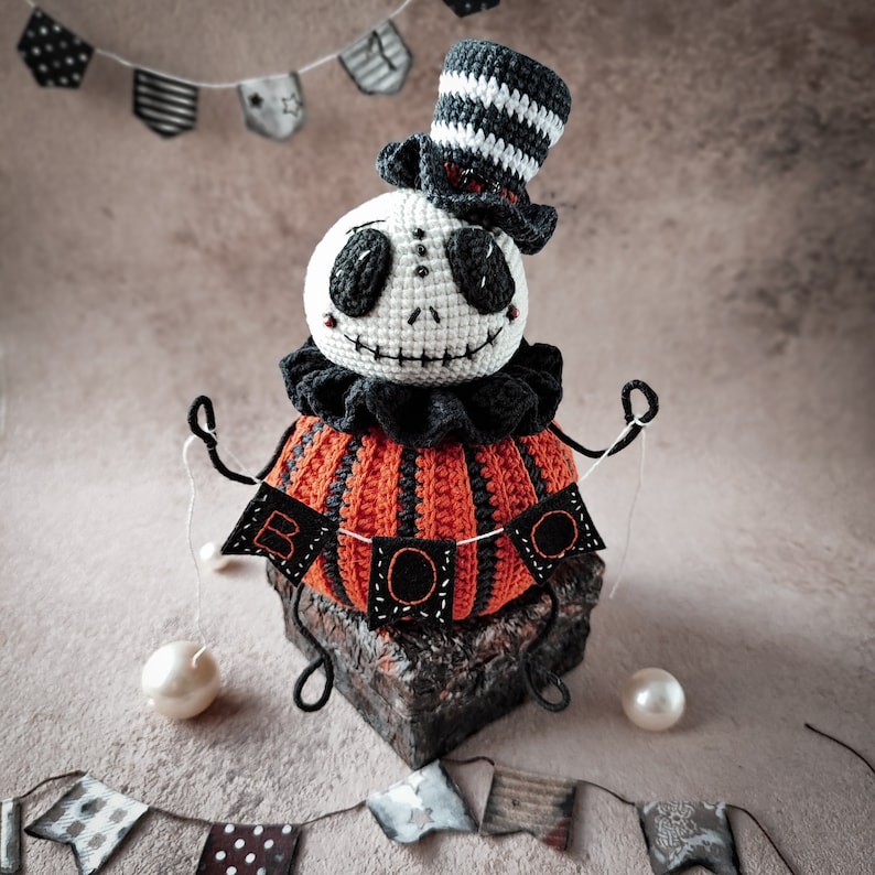 The amigurumi Jack pumpkin toy is displayed on a rustic surface with decorative Halloween elements, including a black "BOO" banner and tiny white pearls. The doll’s curled arms give it a whimsical touch.