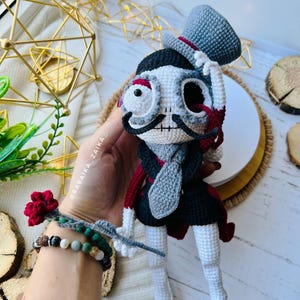 Elegant crochet skeleton doll wearing a black cape and holding a red rose, displayed as a Halloween centerpiece.
Mr. Santa Muerte amigurumi sitting gracefully with a red rose, surrounded by natural wooden textures and greenery.