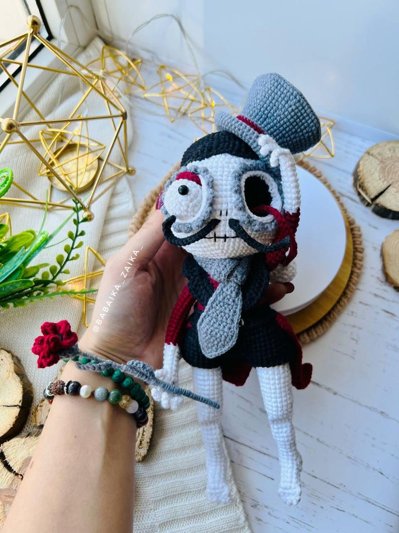 Elegant crochet skeleton doll wearing a black cape and holding a red rose, displayed as a Halloween centerpiece.
Mr. Santa Muerte amigurumi sitting gracefully with a red rose, surrounded by natural wooden textures and greenery.