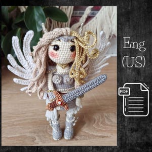 May include: A handmade crochet doll of a warrior with wings, holding a sword. The doll has a light skin tone, blonde hair, and is wearing a gray and gold outfit. The doll is approximately 6 inches tall. The image also includes the text "Eng (US)" and a PDF icon.