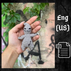 May include: A crocheted gray amigurumi doll of the character,  "Oogie Boogie" from the movie "The Nightmare Before Christmas". The doll has black eyes and a stitched mouth. The doll is being held in a hand.