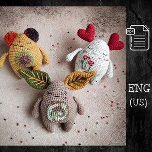 Könnte beinhalten: Drei gehäkelte Amigurumi-Spielzeuge mit verschiedenen Designs. Das erste Spielzeug ist gelb mit einem braunen Gesicht und schwarzen Ohren. Das zweite Spielzeug ist weiß mit roten Herzen und einem Blumendesign. Das dritte Spielzeug ist braun mit grünen und gelben Blättern und einem Wirbeldesign. Die Spielzeuge befinden sich auf einem beigefarbenen Hintergrund mit roten und weißen Perlen.