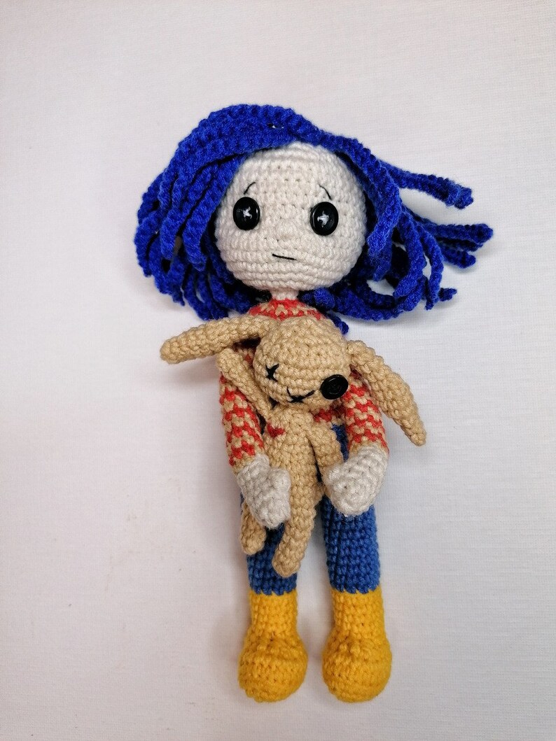 The Coraline doll with its removable raincoat placed beside it, showcasing the outfit customization feature of the crochet pattern.