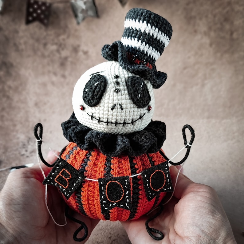 The crochet Halloween pumpkin doll in a festive setting, surrounded by small seasonal decorations. The figure’s arms appear lively and expressive, making it a unique decor piece.