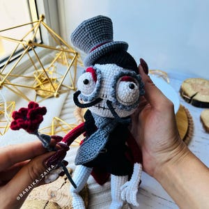 Handmade crochet skeleton doll in black and gray attire, sitting on a wooden display stand with nature-inspired decor.