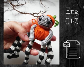 Puede incluir: Araña de crochet con un disfraz de calabaza. La araña tiene patas rayadas en blanco y negro y un cuerpo gris. El disfraz de calabaza es naranja con un tallo verde.