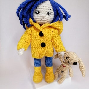 The Coraline doll lying down, with her raincoat removed, revealing a red and white striped sweater and blue crochet pants.
