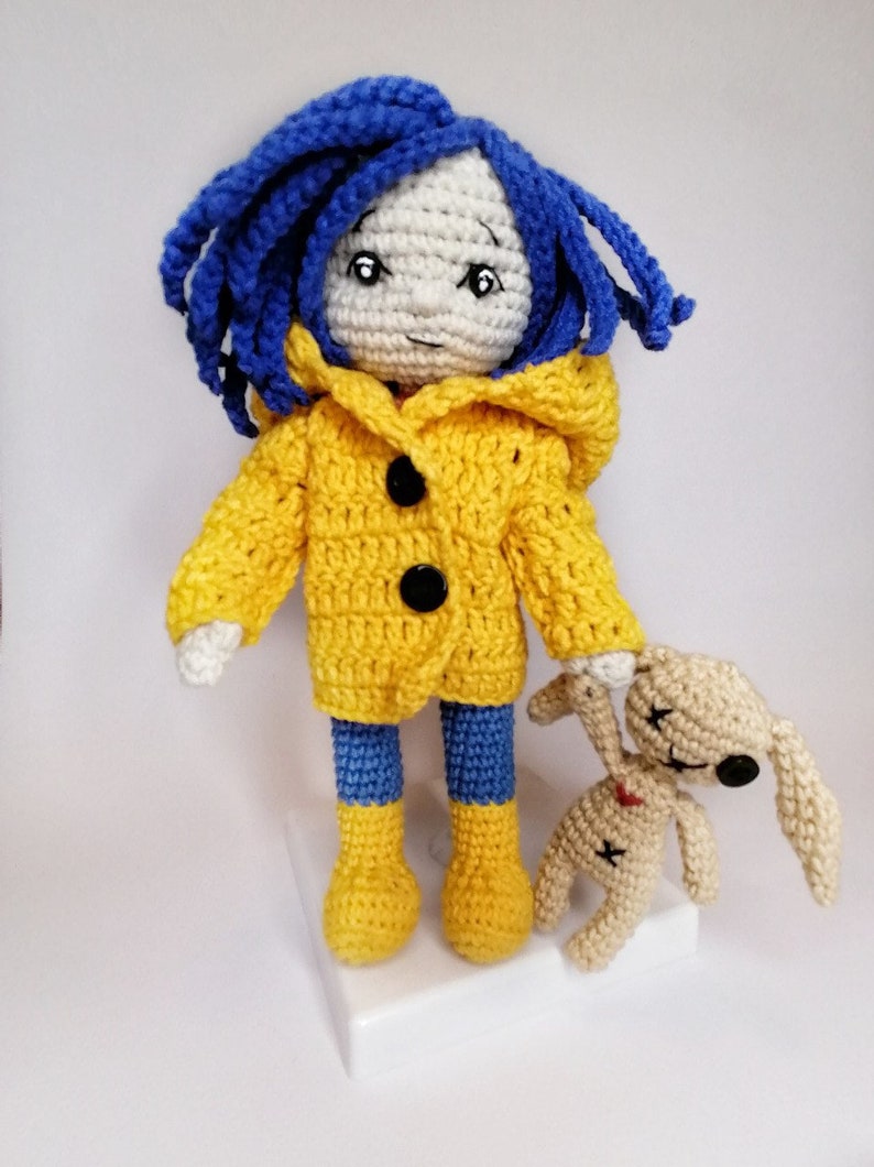 The Coraline doll lying down, with her raincoat removed, revealing a red and white striped sweater and blue crochet pants.