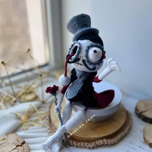 Amigurumi skeleton doll Mr. Santa Muerte with red accents, sitting on a white background with wooden and golden decor.