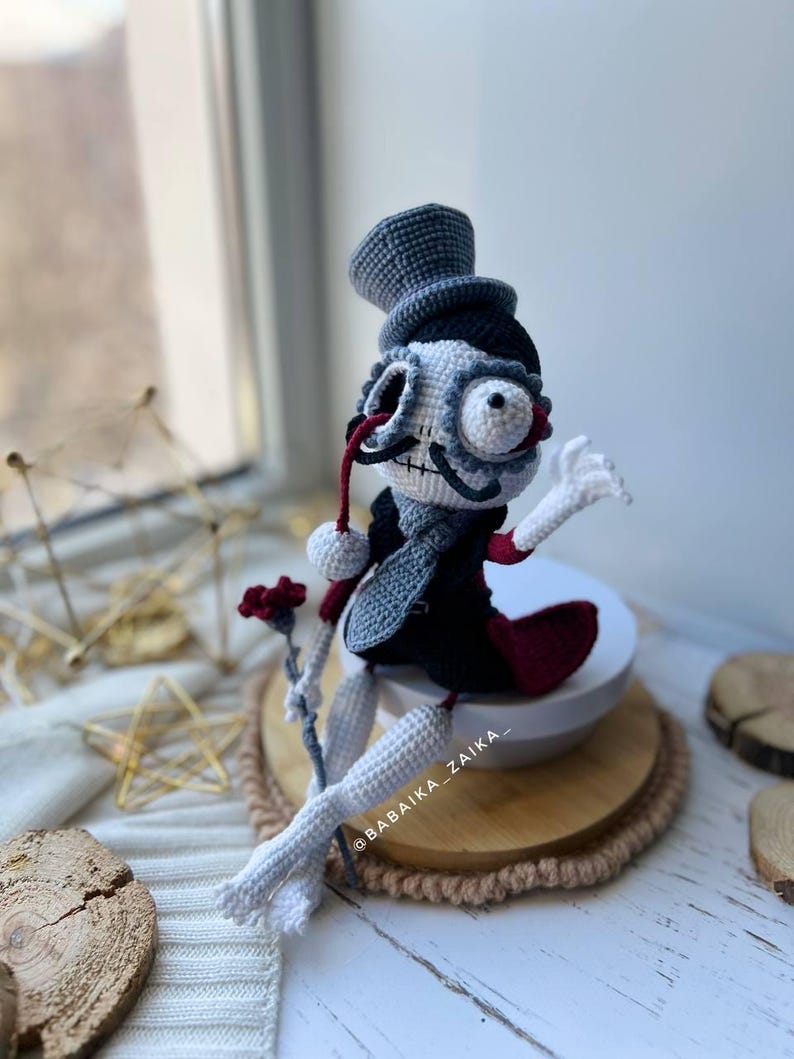 Amigurumi skeleton doll Mr. Santa Muerte with red accents, sitting on a white background with wooden and golden decor.