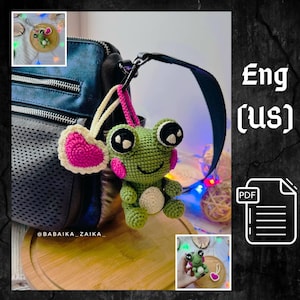 Può includere: Un portachiavi a forma di rana verde all'uncinetto con grandi occhi neri e guance rosa, attaccato a una borsa. Un cuore rosa all'uncinetto con un bordo bianco è appeso accanto alla rana. Il testo "Eng (US)" e un'icona PDF sono visibili.
