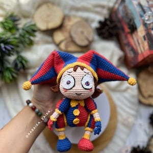 Pomni crochet clown sitting on a white stand, with its detailed red, blue, and yellow design standing out. The perfect addition to your handmade Halloween decor or a quirky collector's item.