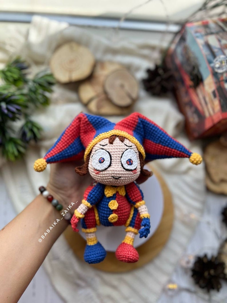 Pomni crochet clown sitting on a white stand, with its detailed red, blue, and yellow design standing out. The perfect addition to your handmade Halloween decor or a quirky collector's item.