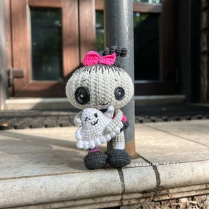 The crochet doll posed against a fence with its ghost buddy, ideal for Halloween-themed decorations. The fine crochet stitches and amigurumi techniques make it a wonderful DIY handmade craft.