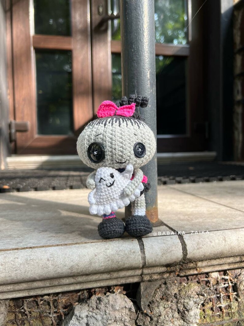 The crochet doll posed against a fence with its ghost buddy, ideal for Halloween-themed decorations. The fine crochet stitches and amigurumi techniques make it a wonderful DIY handmade craft.