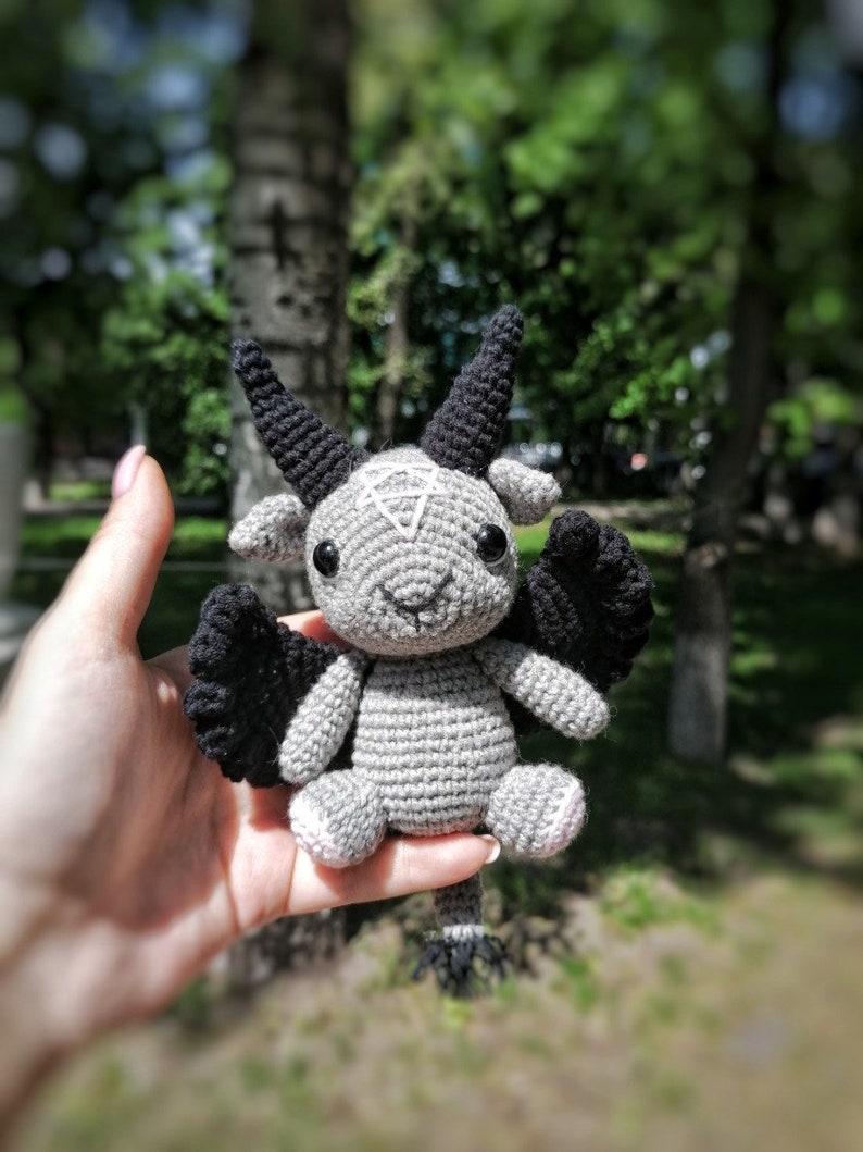 Crocheted Baphomet toy positioned on a rustic outdoor background, showcasing its black wings and mystical charm.