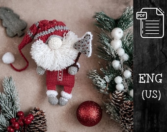 May include: Crocheted gnome toy wearing a red and white hat and holding a red and white axe. The gnome is sitting on a brown surface with a pine branch, pine cones, and red berries in the background. The image also includes a red glitter ball and a white pearl.