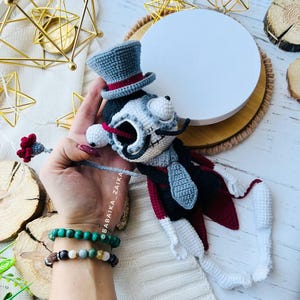 Mr. Santa Muerte crochet doll in a stylish outfit, with a gray top hat and a red cape, positioned against rustic background elements.