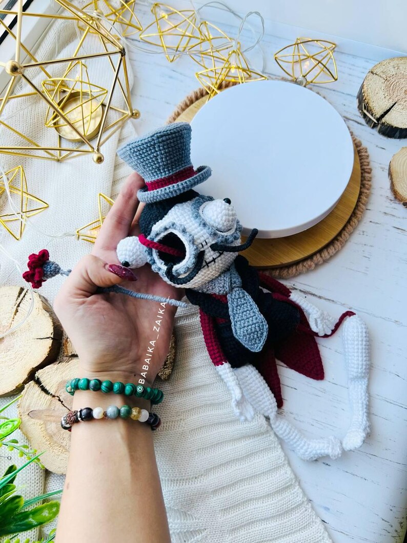 Mr. Santa Muerte crochet doll in a stylish outfit, with a gray top hat and a red cape, positioned against rustic background elements.