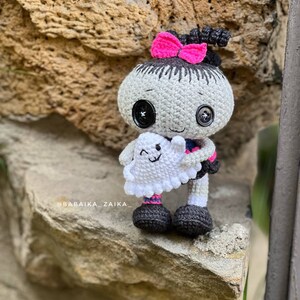 The crochet doll sitting on a ledge outdoors, displaying its full design. The pink accents on the dress and bow contrast beautifully with the black and gray body, making it a unique handmade Halloween decor or gift.