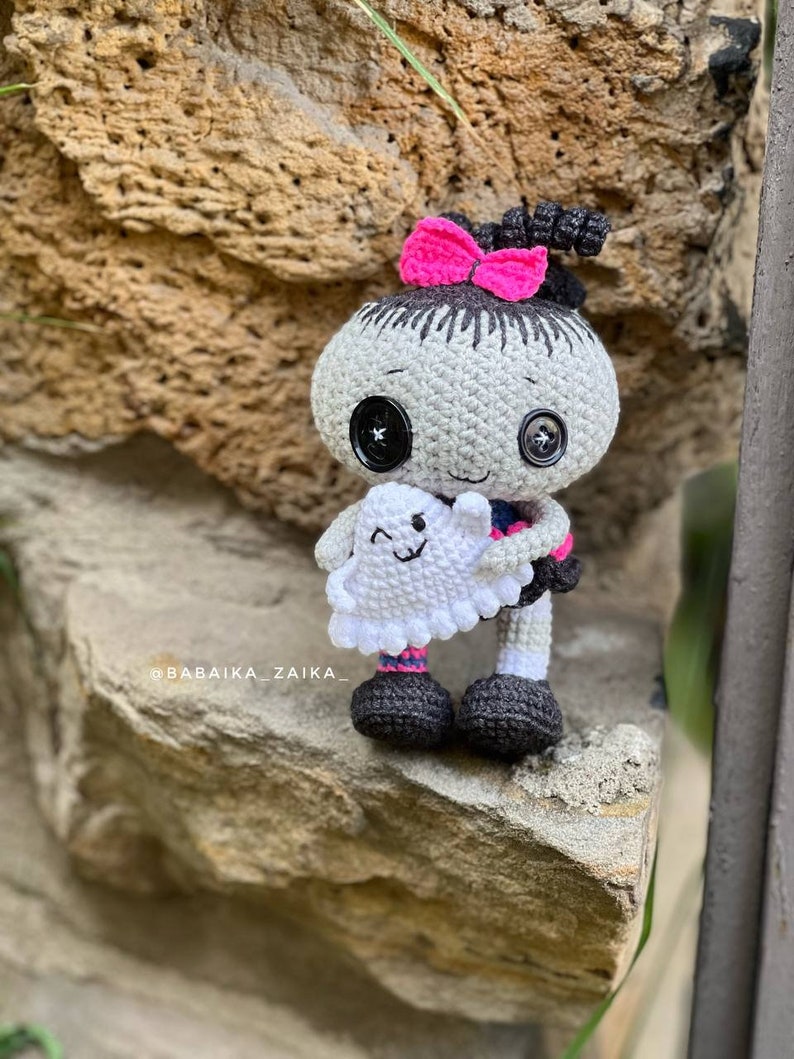 The crochet doll sitting on a ledge outdoors, displaying its full design. The pink accents on the dress and bow contrast beautifully with the black and gray body, making it a unique handmade Halloween decor or gift.