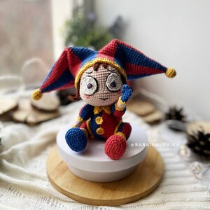Finished Pomni amigurumi clown doll sitting on a wooden base, dressed in a vibrant red and blue crochet outfit with a jester hat. Perfect for Halloween-themed decor or a fun handmade toy gift.