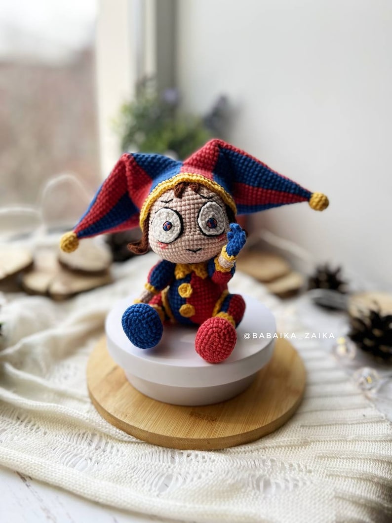 Finished Pomni amigurumi clown doll sitting on a wooden base, dressed in a vibrant red and blue crochet outfit with a jester hat. Perfect for Halloween-themed decor or a fun handmade toy gift.