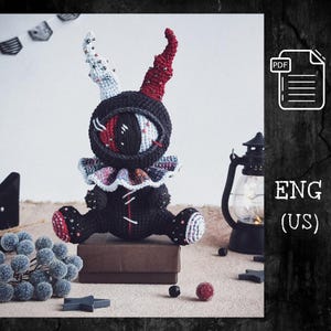 May include: A crocheted plush toy featuring a large central eye, one white horn, one red horn, and a ruffled collar with multicolored accents. The body is black with white stitching details, red accents on the feet, and small metallic embellishments.