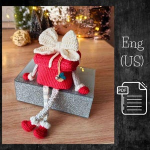 May include: A handmade, red crochet gift box doll with a cream-colored bow, arms, legs, and feet. The doll has a small charm and is sitting on a glittery silver box. The background includes Christmas decorations.