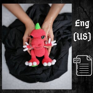 May include: A crocheted pink dinosaur toy with white accents and green spikes. The dinosaur is being held by a person's hands.