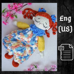 Puede incluir: Patrón de muñeca de crochet en inglés (EE. UU.) en formato PDF. La muñeca está tumbada con los ojos cerrados, tiene el pelo rojo, un cuello azul y un vestido colorido. La muñeca está sobre una superficie blanca con flores rosas.