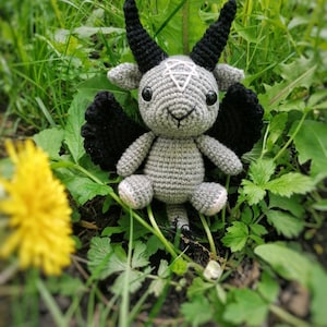 Crocheted Baphomet toy posed near yellow dandelions in the grass, creating a contrast between the dark tones and bright flowers.