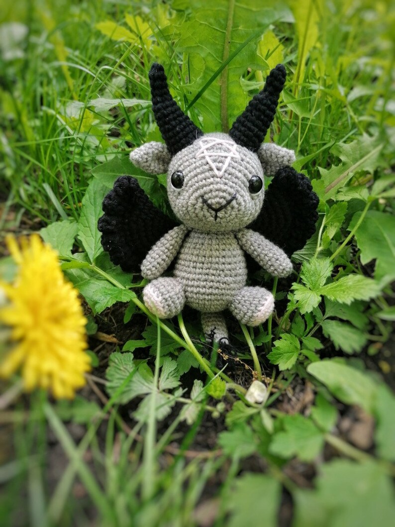 Crocheted Baphomet toy posed near yellow dandelions in the grass, creating a contrast between the dark tones and bright flowers.