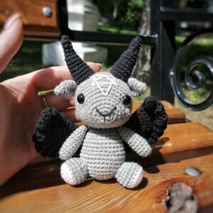 Crocheted Baphomet toy placed on a wooden surface, emphasizing the occult-inspired details like black wings and horns.