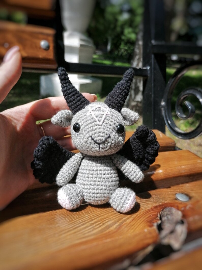 Crocheted Baphomet toy placed on a wooden surface, emphasizing the occult-inspired details like black wings and horns.