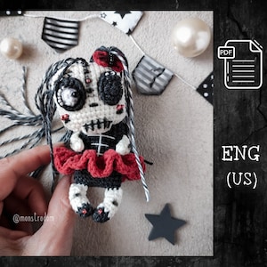 Może przedstawiać: Lalka amigurumi szydełkowa z czaszką, czarnymi włosami, czerwoną i czarną sukienką oraz czerwonymi i czarnymi butami. Lalka trzyma czerwoną kokardkę. Lalka siedzi na dłoni. Tło to szara powierzchnia z czarno-białymi flagami w paski.
