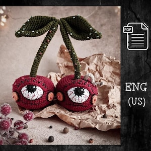 May include: Two crocheted cherries with green stems and leaves. Each cherry has a large, white eye with a black pupil and red veins. The cherries are sitting on a brown paper background.