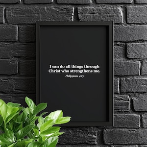 May include: A framed black print with white text that reads, "I can do all things through Christ who strengthens me. Philippians 4:13." The print is set against a black brick wall, with green leafy plants in the lower left corner.