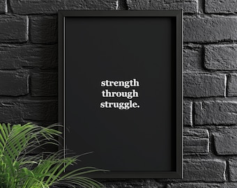 Könnte beinhalten: Schwarzer gerahmter Druck mit dem weißen Text "strength through struggle". Das Kunstwerk ist vor einer schwarzen Backsteinmauer platziert, mit einer grünen Pflanze in der unteren linken Ecke.