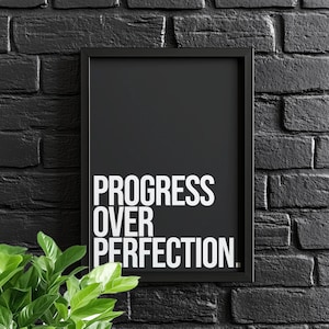 May include: Black framed print with the words "PROGRESS OVER PERFECTION" in bold white letters. The print is against a black brick wall, with green leafy plants in the lower left corner.