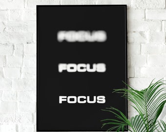 以下が含まれることがあります： 白黒のポスターに「FOCUS」という単語が3回繰り返されています。上の2つはぼやけていて、下の1つはピントが合っています。ポスターは黒の額縁で囲まれ、白いレンガの壁に掛けられています。