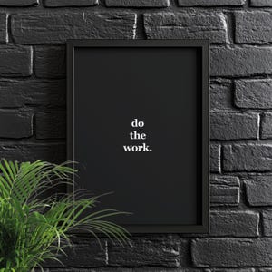 May include: Black and white motivational poster with a black frame hanging on a black brick wall. The poster says "do the work."