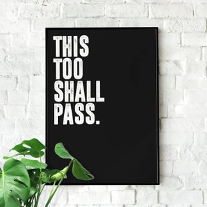 May include: Black and white framed poster with the text "THIS TOO SHALL PASS." hanging on a white brick wall.