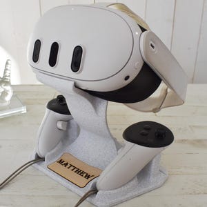 Op de afbeelding: Een witte VR-headset en controllers rusten op een grijze gespikkelde standaard. De standaard heeft een houten naamlabel met de tekst "MATTHEW". De VR-headset heeft drie zwarte ventilatieopeningen en een zwart vizier. De controllers zijn wit met zwarte knoppen.
