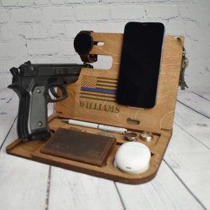 Może przedstawiać: Drewniany organizer na biurko z przegr&oacute;dkami na telefon, zegarek, portfel, pierścionki i słuchawki. Posiada wycinany laserowo wz&oacute;r amerykańskiej flagi z niebieską linią i nazwiskiem "WILLIAMS". Czarny pistolet umieszczono po lewej stronie.