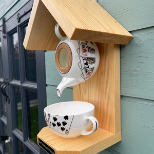 May include: A wooden bird feeder shaped like a house with a teapot pouring into a teacup. The teapot has a playing card design and the teacup has a black spade design. The feeder has a sign that reads "Down To Earth Garden Features".