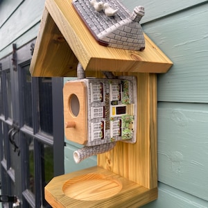 May include: A wooden bird feeder featuring a ceramic teapot shaped like a building with the text "HIGHWOOD PACK" on the side. The feeder has a gabled roof topped with two small white bird figurines, a circular entrance hole, and a wooden base tray.