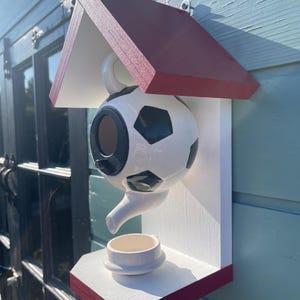 May include: A novelty birdhouse in the shape of a football, with a red roof and a white base. The birdhouse has a small cup-shaped perch and is mounted on a light blue wooden surface.